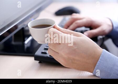 Mann trinkt Kaffee, der an der PC-Tastatur sitzt. Tasse heißes Getränk in der männlichen Hand, Pause während der Arbeit im Büro Stockfoto