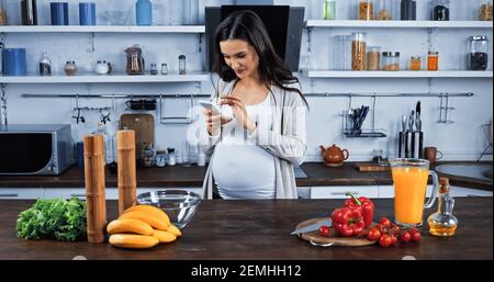 Schwangere Frau mit Smartphone in der Nähe von Rohstoffen in der Küche Stockfoto