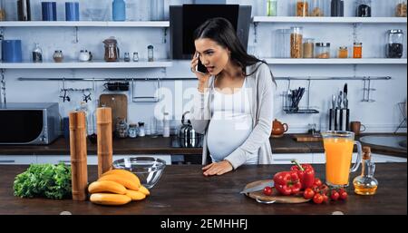 Schwangere Frau im Gespräch auf dem Smartphone in der Nähe von frischen Lebensmitteln in der Küche Stockfoto