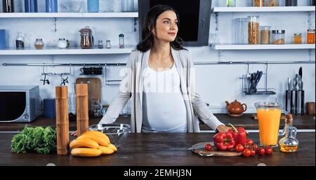 Schwangere Frau steht in der Nähe von Bio-Lebensmittel und Orangensaft auf Küchentisch Stockfoto