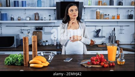 Nachdenkliche schwangere Frau in der Nähe von Smartphone und frische Zutaten in Küche Stockfoto