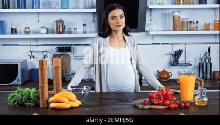 Schwangere Frau, die Kamera in der Nähe von frischem Gemüse und Orange Saft Stockfoto