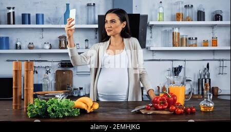 Schwangere Frau, die Selfie in der Nähe von Zutaten auf Küchentisch Stockfoto