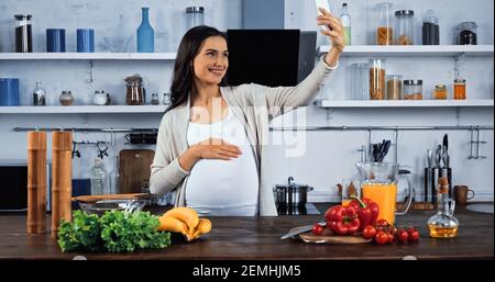 Lächelnde schwangere Frau, die Selfie in der Nähe von frischem Essen in der Küche Stockfoto