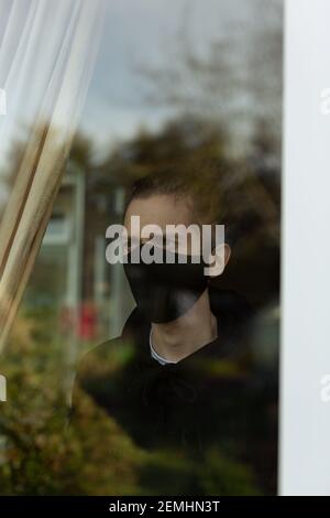 Ein junger Mann mit Gesichtsmaske, der während seiner Selbstisolation zu Hause aus dem Fenster schaut. Covid-19 Corona Virus Isolation Einsamkeit Konzept Stockfoto