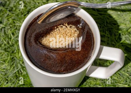 Ein Teelöffel gießt braunen Zucker in eine Tasse Kaffee, Nahaufnahme. Süße das Getränk mit Rohrzucker. Stockfoto