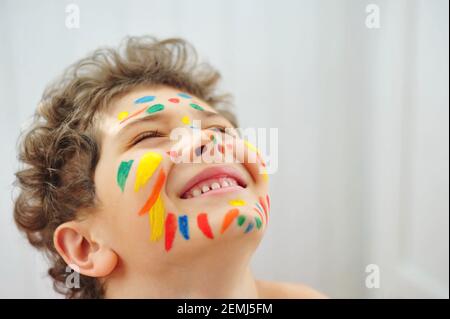 Ein süßer kleiner Junge malte bunte Streifen auf seinem Gesicht mit Farben. Glückliches Kind denkt, dass er ein Clown oder ein echter Inder ist. Kind lächelt. Stockfoto