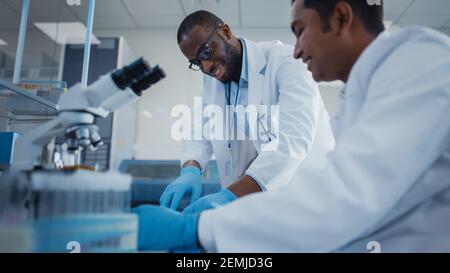 Modernes Medizinisches Forschungslabor: Zwei Lächelnde Männliche Wissenschaftler Arbeiten Mit Mikroskop Zusammen, Analysieren Proben, Sprechen. Erweitertes Wissenschaftliches Labor Stockfoto