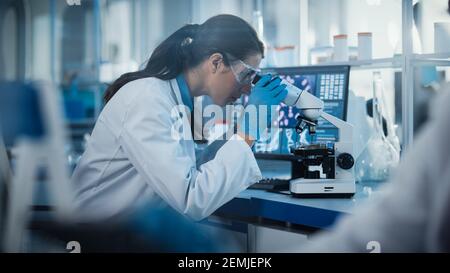 Medical Research Laboratory: Portrait einer schönen Wissenschaftlerin in Goggles schaut unter Mikroskop auf Proben. Advanced Scientific Lab für Stockfoto