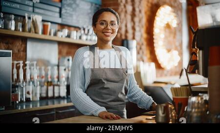 Schöne lateinamerikanische weibliche Barista mit kurzen Haaren und Gläsern projiziert ein Happy Smile in Coffee Shop Bar. Portrait des glücklichen Mitarbeiters dahinter Stockfoto