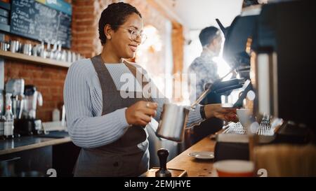 Schöne lateinamerikanische weibliche Barista mit kurzen Haaren und Gläsern macht eine Tasse leckeren Cappuccino in Coffee Shop Bar. Porträt eines glücklichen Mitarbeiters Stockfoto