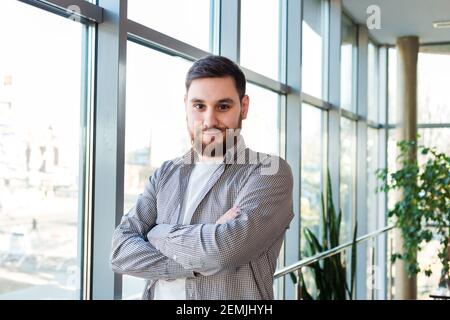 Selbstbewusster kaukasischer Mann, der in der Nähe des großen Fensters im Büro mit Pflanzen steht. Erfolgreicher bärtiger junger Mann kreuzte die Arme. Gut aussehender Geschäftsmann Stockfoto