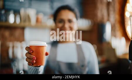 Schöne lateinamerikanische weibliche Barista mit kurzen Haaren und Gläsern lächelt und hält eine Take Away Kaffeetasse in einem Café. Nahaufnahme mit Stockfoto