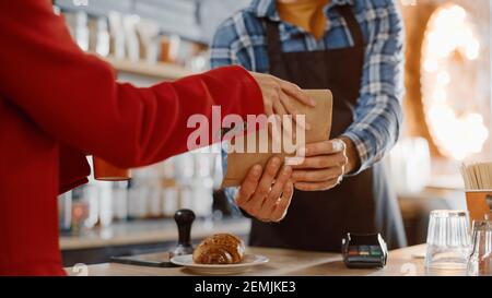 Nahaufnahme Aufnahme einer femininen Hand in Red Coat Taking Paper Bag mit Gebäck und Karton-Tasse mit Take Away Coffee in a Cafe. Happy Employee Works Stockfoto