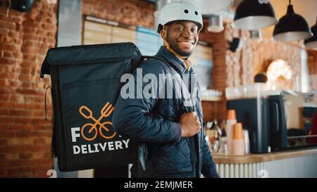 Gutaussehende schwarz afroamerikanische Lebensmittel Lieferung Kurier posiert vor der Kamera in einem Café. Glücklicher und lächelnder Mann, der einen Fahrradhelm trägt Stockfoto