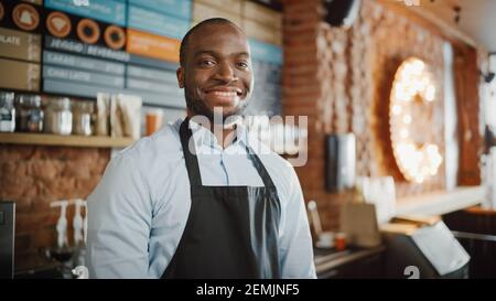 Im Coffee Shop Restaurant lächelt der gutaussehende schwarzafroamerikanische Barista mit kurzen Haaren und Bartschürze. Porträt eines glücklichen Mitarbeiters Stockfoto