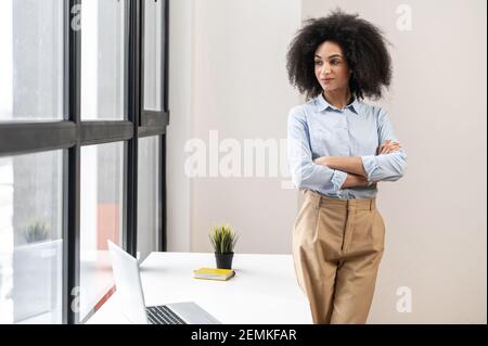 Erfolgreiche selbstbewusste junge Mixed-Race-Unternehmerin oder eine Geschäftsfrau mit Afro-Frisur in legerer Kleidung stehend mit Armen im Büro gefaltet, Blick in das Fenster mit Panoramablick Stockfoto