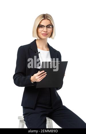 Junger Nachrichtenanker mit Zwischenablage lächelt beim Sitzen an der Kamera Auf Stuhl isoliert auf weiß Stockfoto