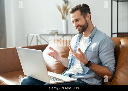 Lächelnder junger bärtiger Hipster, der auf der Couch mit Laptop auf dem Schoß sitzt, flirtet, Dating-App benutzt, Video-anrufende Freundin online, gestikiert und gesprochen, Blogger streamen seinen Alltag Stockfoto