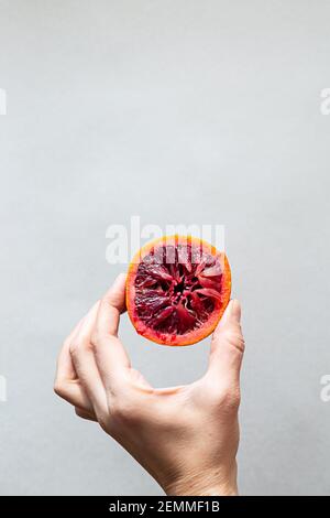 Das moderne Konzept der gesunden Ernährung. Eine weibliche Hand hält die Hälfte einer roten orange Frucht. Minimalistische Komposition auf hellgrauem Hintergrund mit Copy-SPAC Stockfoto