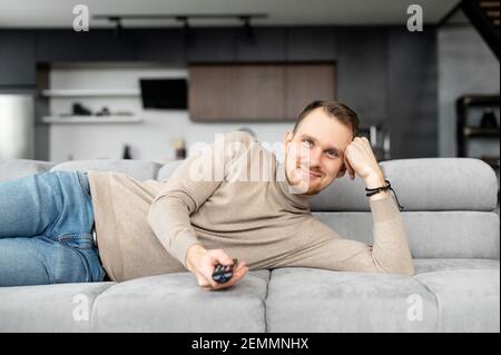 Junger bärtiger Hipster, der auf der Couch im Wohnzimmer liegt, mit einer fernbedienung auf die Kamera zeigt, die Kanäle wechselt, mit einem Fernsehprogramm zufrieden ist, Film guckt, Serien schaut, zu Hause chillt Stockfoto