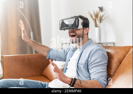 Glücklich aufgeregt jungen bärtigen Hipster Kerl sitzt auf dem Sofa im Wohnzimmer, bekommen Erfahrung der virtuellen Realität zu Hause, trägt VR-Brille oder ein Headset, während mit den Händen im Cyberspace drücken Stockfoto