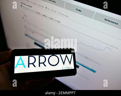 Person, die Mobiltelefon mit Logo der Investmentgesellschaft Arrow Global Group PLC auf dem Bildschirm vor der Webseite mit Diagramm hält. Konzentrieren Sie sich auf die Telefonanzeige. Stockfoto