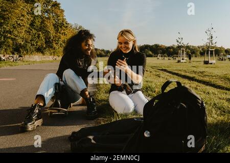 Lächelnde Freundinnen mit Smartphone sitzen im Park Stockfoto
