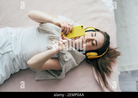 Junge, sorglose Millennials-Frau in Kopfhörern, die auf dem Bett liegt, kommuniziert am Telefon und lächelt. Stockfoto