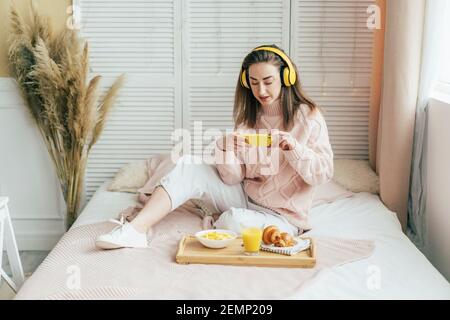 Millennial Mädchen auf dem Bett im Schlafzimmer sitzt macht ein Foto von Frühstück auf einem Tablett hören Musik auf Kopfhörer. Stockfoto