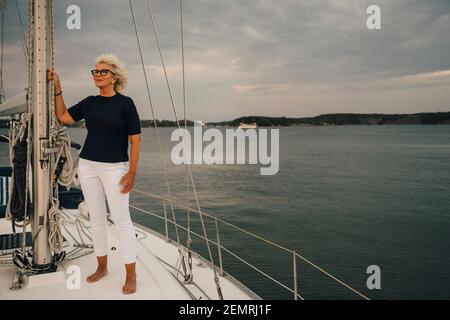 Ältere Frau schaut weg, während sie im Boot gegen den Himmel steht Und Meer bei Sonnenuntergang Stockfoto