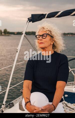 Ältere Frau mit geschlossenen Augen sitzt auf dem Boot während des Sonnenuntergangs Stockfoto