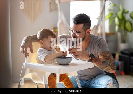 Single Papa Fütterung sein Baby, das in einem gesessen wird Kinderstuhl Stockfoto