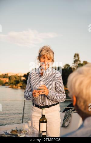 Lächelnde ältere Frau mit Weinglas und Blick auf den Mann im Boot Gegen den Himmel Stockfoto