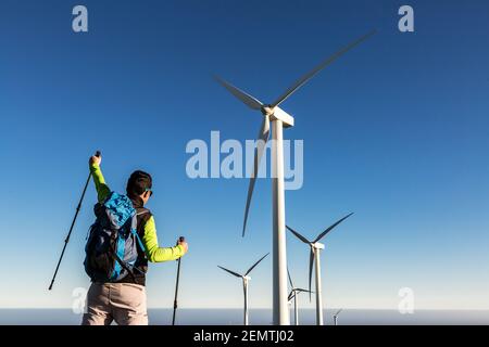 Rückansicht des gesichtslosen männlichen Wanderers mit Trekkingstöcken zum Feiern Triumph mit erhobenem Arm, während er auf dem Hintergrund von Windmühlen steht An sonnigen Tagen auf La Stockfoto