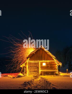 Fantastische Winterlandschaft mit glühender Holzhütte im verschneiten Wald. Feuerfunken fliegen aus dem Kamin. Weihnachtsfeiertagskonzept Stockfoto
