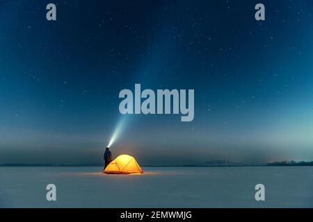 Tourist mit Taschenlampe in der Nähe von gelben Zelt von innen vor dem Hintergrund der unglaublichen Sternenhimmel beleuchtet. Tolle Nachtlandschaft. Touristen Camp in schneebedeckten Feld. Reisekonzept Stockfoto