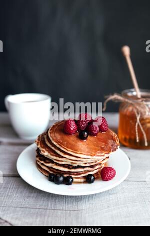 Saftige Pfannkuchen mit Beeren und Honig auf einem weißen Teller, Löffel, Glas, Holztisch, Kaffeetasse. Hochwertige Fotos Stockfoto