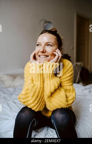 Lächelnde, nachdenkliche Frau, die wegschaut, während sie im Schlafzimmer sitzt Stockfoto