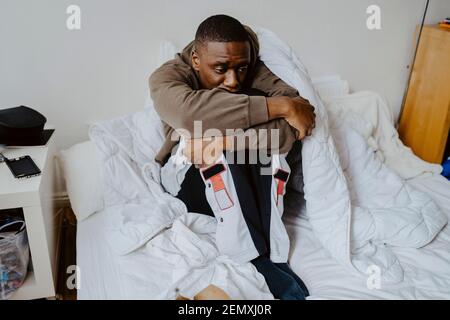 Besorgt Mann umarmt Knie, während auf dem Bett zu Hause sitzen Stockfoto