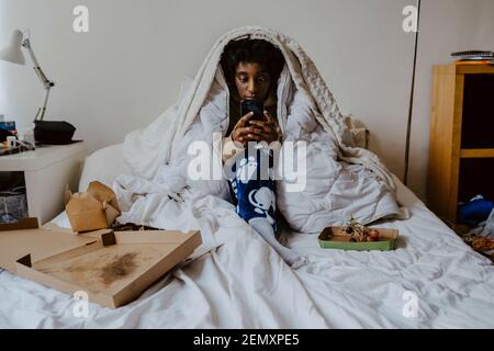 Junge Frau mit Smartphone, während sie im Schlafzimmer sitzt Stockfoto