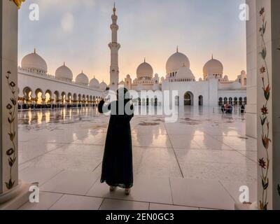 Eine junge muslimische Frau, die einen schwarzen Abaya trägt, macht ein Selfie in einer sehr schönen Moschee bei Sonnenuntergang in Abu Dhabi, VAE. Stockfoto