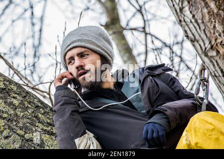 London, Großbritannien. 23. Februar 2021 Der Umweltaktivist Marcus Decker, 32, kommuniziert telefonisch mit seinen Unterstützern. Es ist der zweite Tag des „Baumpflegesitzens“ in den Londoner York Gardens, Battersea. Um einen 100 Jahre alten schwarzen Pappelbaum vor dem Fällen zu retten, sind drei Aktivisten in der Nacht zwischen dem 21. Und 22. Februar geklettert und besetzt. Der Baum sollte gemäht werden, um Platz für ein neues Elektrokabel zu schaffen, Teil des lokalen Wohnungserneuerungsprogramms von council und Taylor Wimpey Homes. Stockfoto