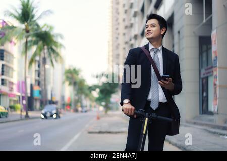Asiatische Geschäftsmann ist Reiten ein Elektroroller und mit seinem Mobiltelefon, um eine Karte in der Anwendung zu öffnen und zu prüfen Die Stadtstrassen Routen in der m zu arbeiten Stockfoto