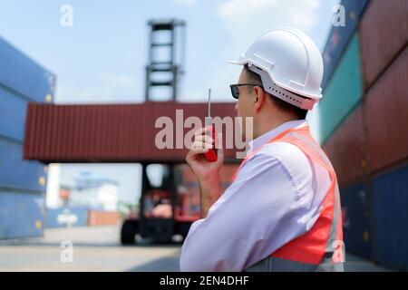 Kaukasischer Vorarbeiter Mann verwendet Walkie Talkie, um seine Arbeiter mit Kran heben Container im Hintergrund in Container Depot Terminal Befehl. Stockfoto