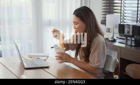 Asiatische Geschäftsfrau essen Instant Nudeln während der Arbeit auf Laptop-Computer zu Hause Büro, glücklich schöne junge Frau sitzen auf Schreibtisch Arbeit overtim Stockfoto