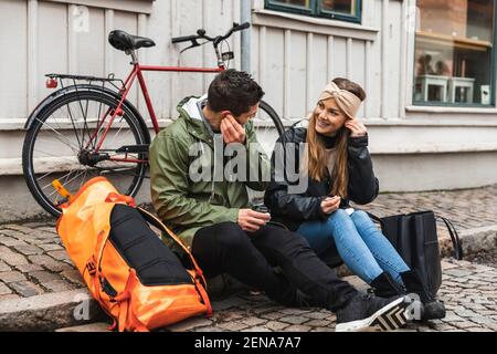 Männliche und weibliche Touristen tragen in-Ear-Kopfhörer, während sie sitzen Fußweg in der Stadt Stockfoto