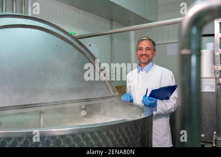 Lebensmittelingenieur in weißer Uniform und blauen Handschuhen mit Blick auf die Kamera. Pasteurisierung der Milch in der Verarbeitungsanlage für Milchprodukte. Lebensmittelsicherheit In Molkereianlagen. Stockfoto