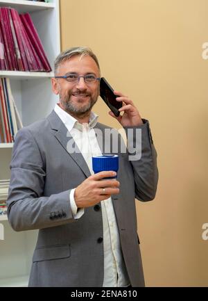 Handsome Business Executive Mit Smartphone. Mann im Anzug hält eine Tasse Kaffee und schaut auf die Kamera. Stockfoto