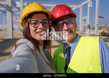 Lächelnder Mann und Frau in Schutzkleidung fotografieren sich mit einer Handykamera vor Ort. Architektin und Bauingenieurin. Stockfoto